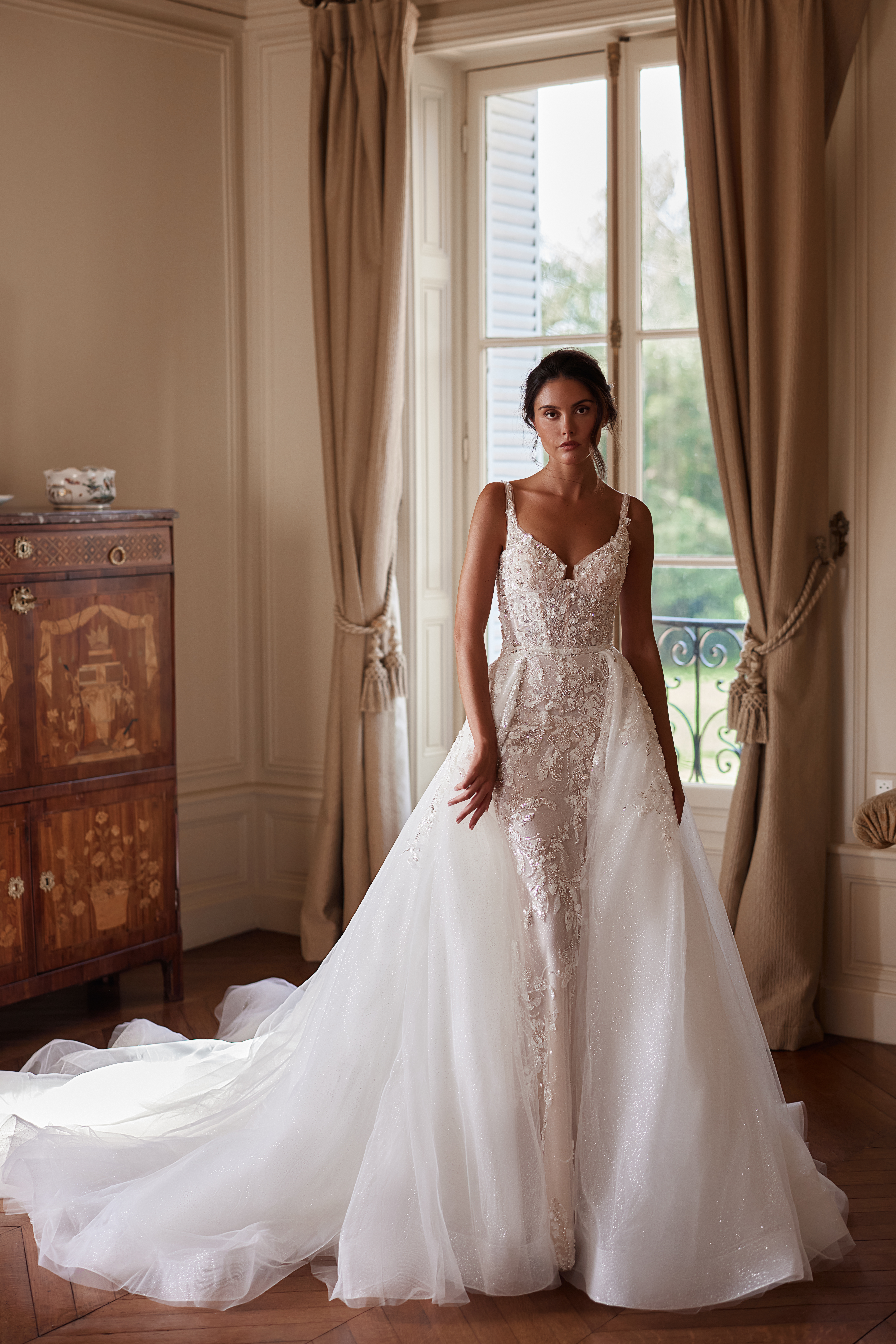 Свадебное платье с кружевными деталями и прозрачной юбкой, модель стоит перед окном в интерьере с деревянной мебелью.