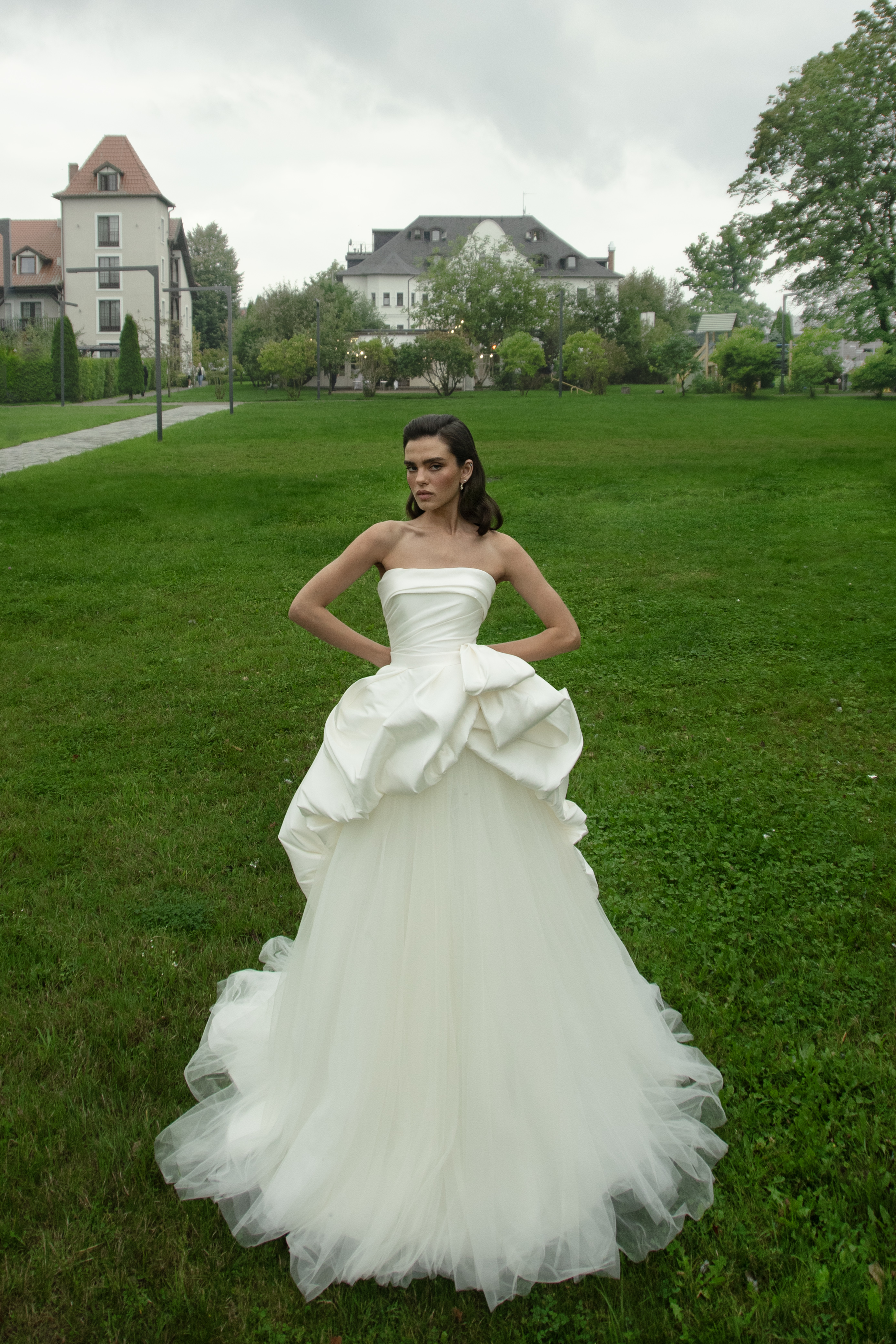 Модель в белом свадебном платье с корсетом и драпировкой позирует на зелёной лужайке на фоне здания.