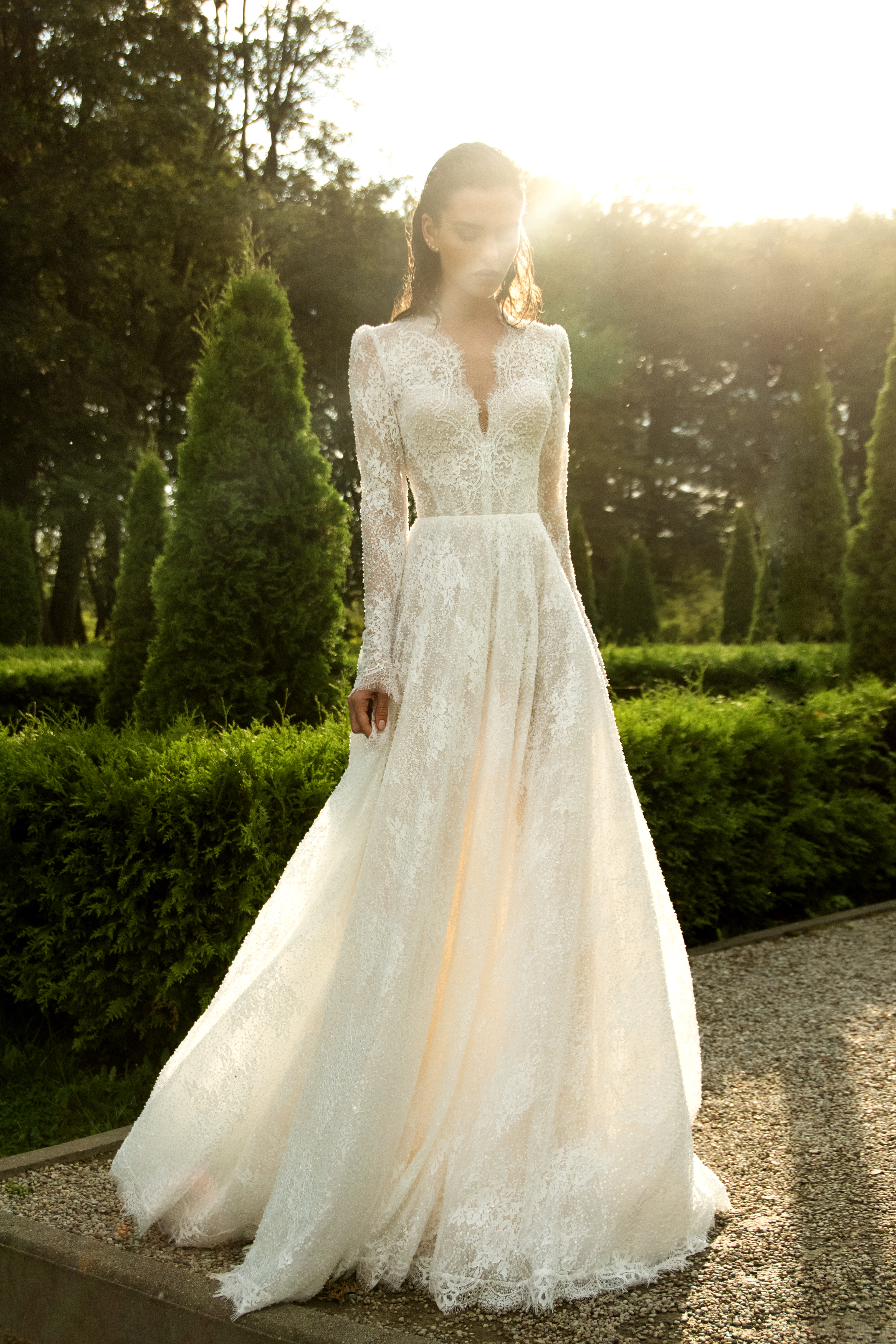 Модель в свадебном платье из кружева с длинными рукавами, стоит на фоне зелёных кустарников и солнечного света.